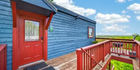 Exterior view of a blue cabin with a red door and deck overlooking a wide green landscape.