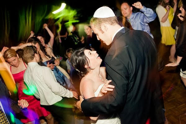 A couple dancing at a wedding reception with guests on a dance floor under colored lighting.