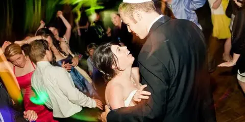 A couple dancing at a wedding reception with guests on a dance floor under colored lighting.