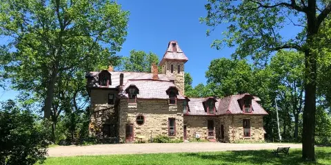 Exterior view of a historic stone castle with a tower and red roof surrounded by green trees and a lawn.