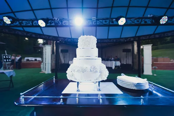 A multi-tiered white wedding cake on a table in a reception venue with stage lighting and a DJ setup in the background.