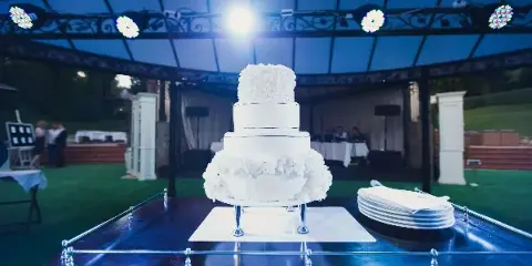 A multi-tiered white wedding cake on a table in a reception venue with stage lighting and a DJ setup in the background.