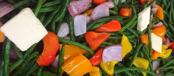 A prepared catering dish consisting of sautéed green beans, red and yellow bell peppers, red onions, and cubes of white cheese.