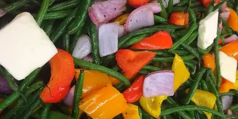 A prepared catering dish consisting of sautéed green beans, red and yellow bell peppers, red onions, and cubes of white cheese.