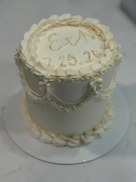 A two-tier white wedding cake decorated with elaborate cream piping, pearl accents, and gold lettering on top.
