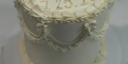 A two-tier white wedding cake decorated with elaborate cream piping, pearl accents, and gold lettering on top.