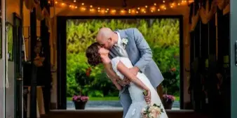 A bride and groom sharing a dip in a wedding venue decorated with string lights, with a garden visible through the open doorway.