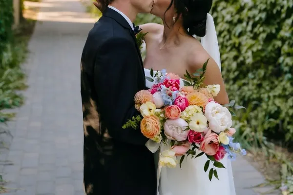 A bride in a white wedding dress holding a vibrant bouquet of pink, orange, and white flowers while kissing a groom in a black suit.