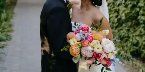 A bride in a white wedding dress holding a vibrant bouquet of pink, orange, and white flowers while kissing a groom in a black suit.