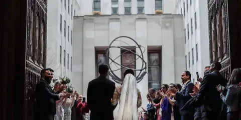 A bride and groom walking away from the camera through a line of applauding wedding guests in an urban courtyard.