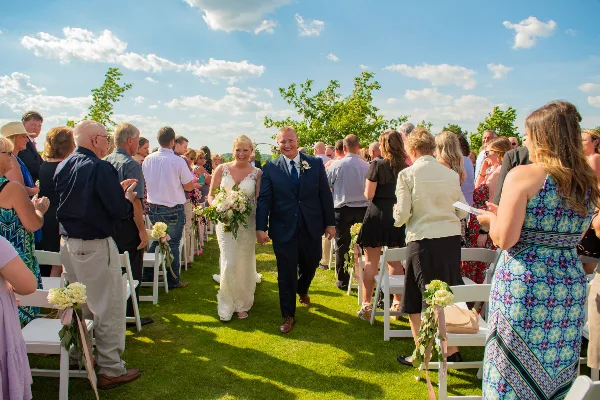 A bride and groom walking down the aisle during an outdoor wedding ceremony, surrounded by seated guests.