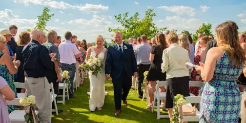 A bride and groom walking down the aisle during an outdoor wedding ceremony, surrounded by seated guests.