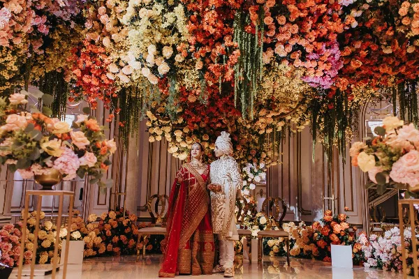 A bride and groom in traditional Indian wedding attire standing in front of a large, elaborate floral installation.