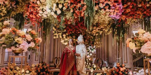 A bride and groom in traditional Indian wedding attire standing in front of a large, elaborate floral installation.