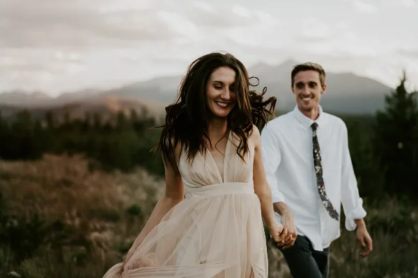 A bride and groom holding hands and smiling while walking through a grassy field with mountains in the background.