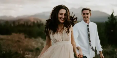 A bride and groom holding hands and smiling while walking through a grassy field with mountains in the background.