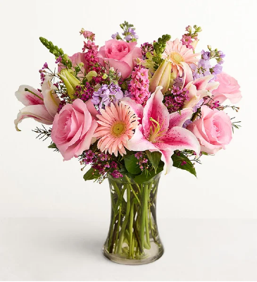 A floral bouquet featuring pink roses, lilies, and purple filler flowers arranged in a clear glass vase.
