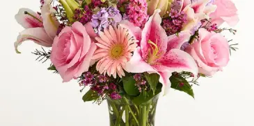 A floral bouquet featuring pink roses, lilies, and purple filler flowers arranged in a clear glass vase.