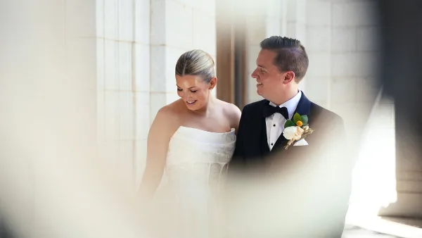 A cinematic wedding film still showing a bride in a white gown and a groom in a tuxedo standing together in a bright, architectural setting.