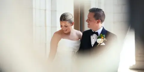 A cinematic wedding film still showing a bride in a white gown and a groom in a tuxedo standing together in a bright, architectural setting.