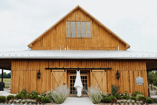 Exterior of a large wooden barn with a wedding dress hanging on the front door.