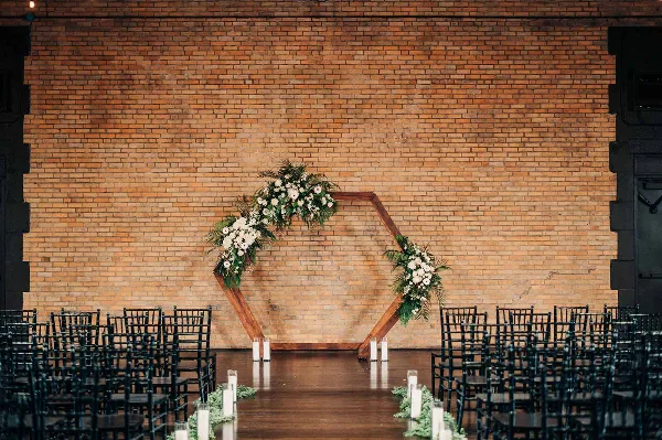 A wedding ceremony setup with a floral arch and rows of black chairs against a large exposed brick wall.