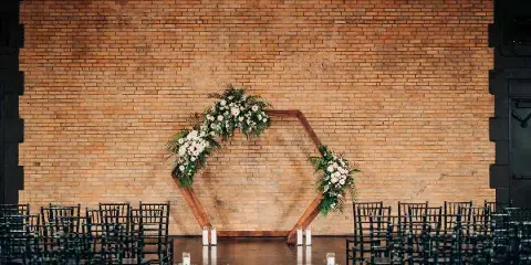 A wedding ceremony setup with a floral arch and rows of black chairs against a large exposed brick wall.