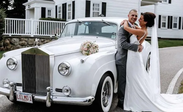 A bride and groom posing next to a white vintage Rolls-Royce with a Le Limo license plate and floral decoration.