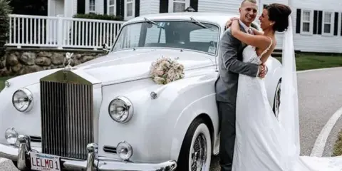 A bride and groom posing next to a white vintage Rolls-Royce with a Le Limo license plate and floral decoration.