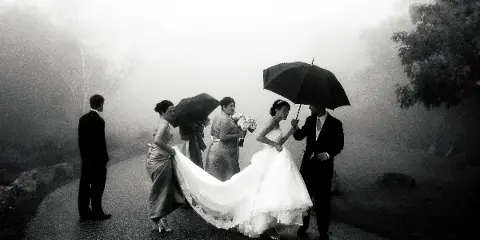 A black-and-white photograph of a bride and groom with bridesmaids holding umbrellas on a foggy path.
