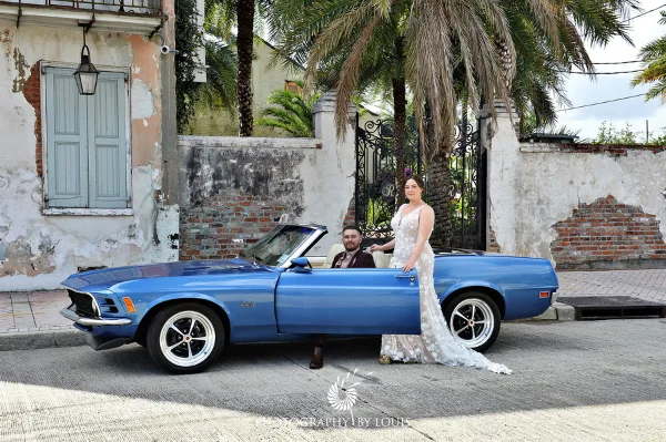 A bride and groom posing with a blue classic convertible car in front of a historic building.