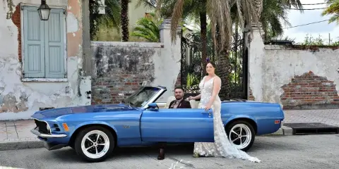 A bride and groom posing with a blue classic convertible car in front of a historic building.