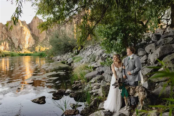 A bride and groom standing with their dog by a river in a rocky, mountainous landscape.