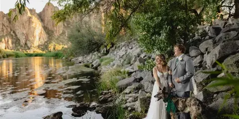 A bride and groom standing with their dog by a river in a rocky, mountainous landscape.