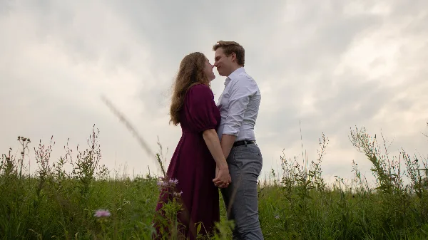 A couple standing in a grassy field holding hands and looking at each other under a cloudy sky.