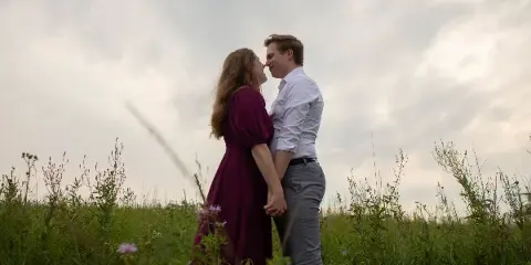 A couple standing in a grassy field holding hands and looking at each other under a cloudy sky.
