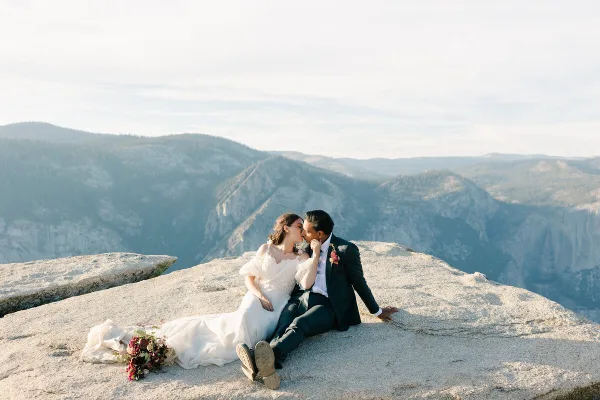 A bride and groom sitting on a rocky mountain ledge with a scenic valley background.