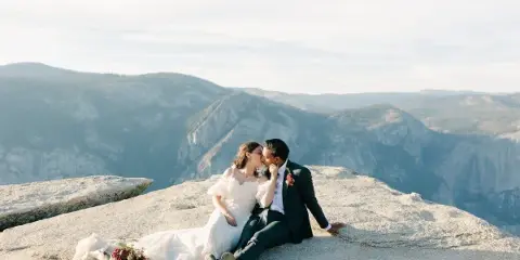 A bride and groom sitting on a rocky mountain ledge with a scenic valley background.