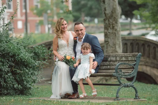 A bride, groom, and young child sitting together on a park bench in formal wedding attire.