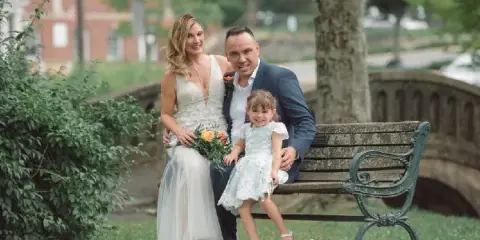 A bride, groom, and young child sitting together on a park bench in formal wedding attire.