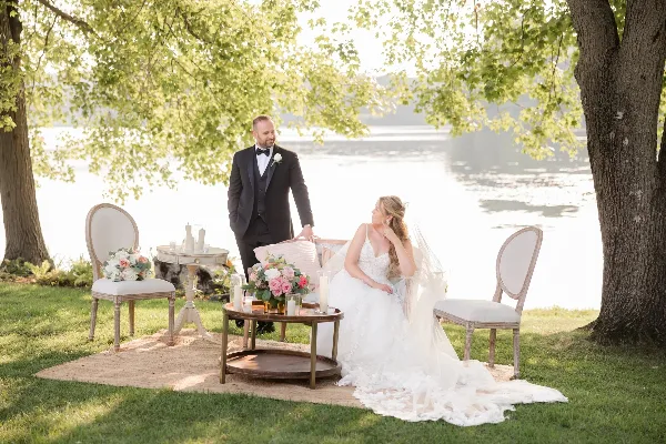 A bride and groom seated on a vintage loveseat and chairs in an outdoor garden setting, surrounded by floral arrangements and candles.