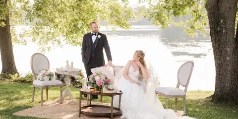 A bride and groom seated on a vintage loveseat and chairs in an outdoor garden setting, surrounded by floral arrangements and candles.