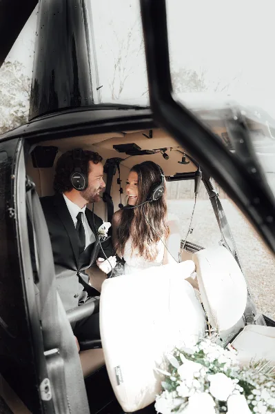 A bride and groom in wedding attire sitting inside a helicopter, smiling at each other.