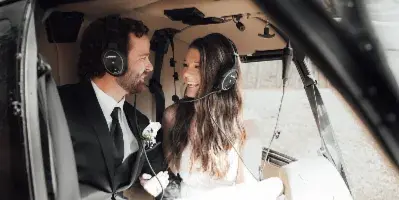 A bride and groom in wedding attire sitting inside a helicopter, smiling at each other.