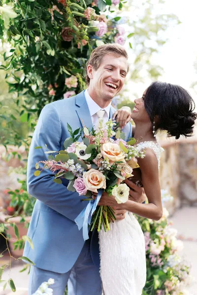 A bride and groom laughing together under a floral arch, with the bride holding a large bouquet.