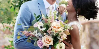 A bride and groom laughing together under a floral arch, with the bride holding a large bouquet.