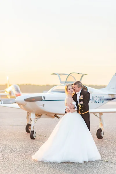 Bride in a white gown and groom in a military dress uniform embracing in front of a small airplane at sunset.
