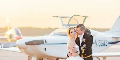Bride in a white gown and groom in a military dress uniform embracing in front of a small airplane at sunset.