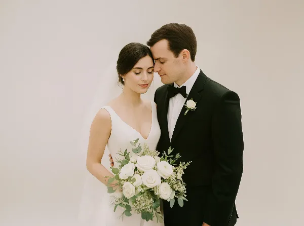 A bride in a white gown and a groom in a black tuxedo standing together holding a white rose bouquet.