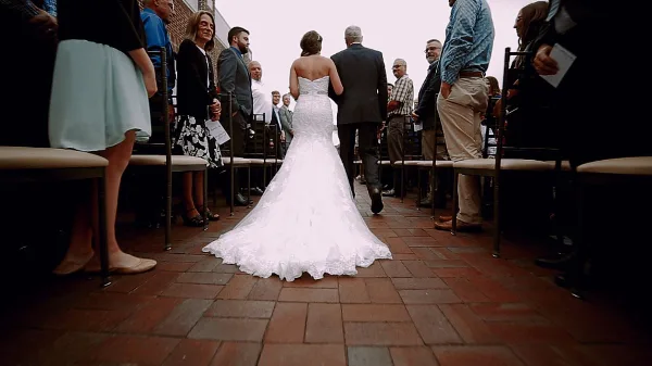 A bride in a white lace gown walking down a brick aisle toward a wedding ceremony with guests seated on either side.